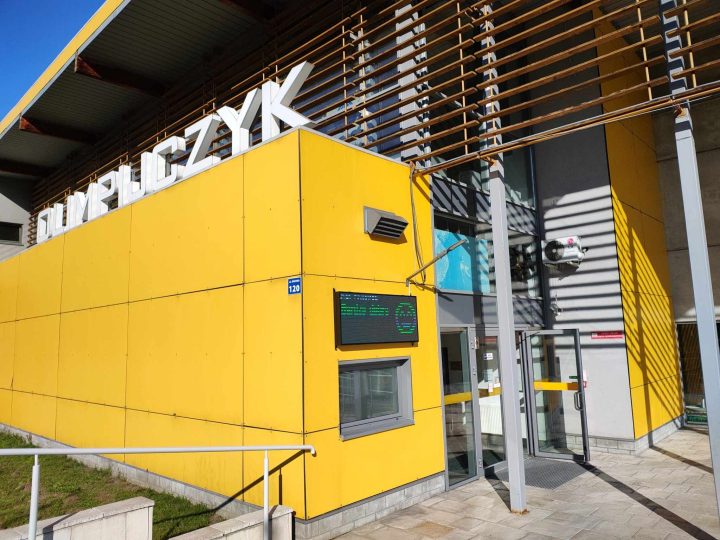 A modern building with a bright yellow exterior, featuring large letters on top and a digital display near the entrance.