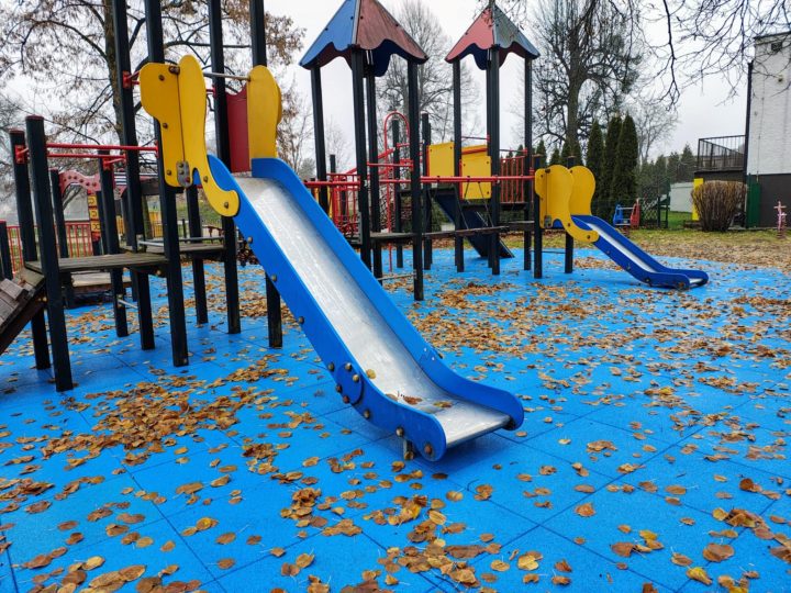 A playground featuring two blue slides, surrounded by fallen leaves on a blue rubber surface.