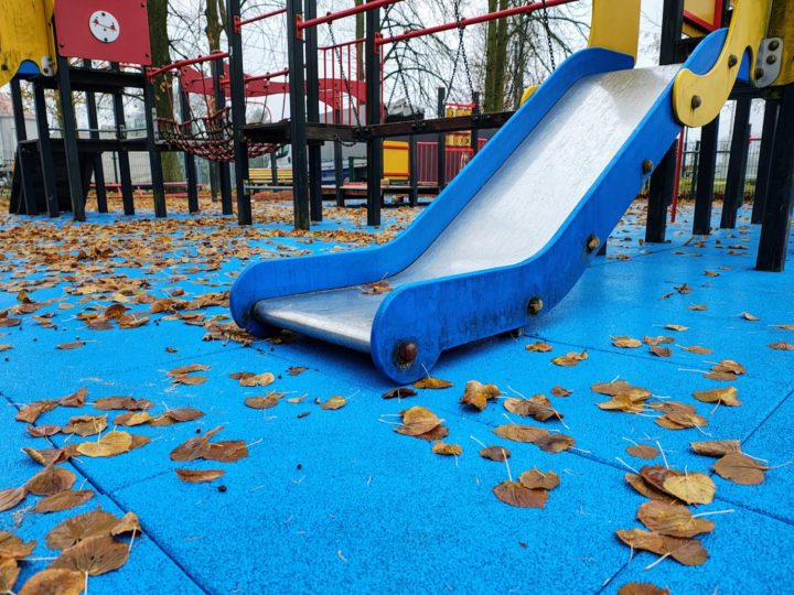 A playground slide on a blue surface, surrounded by fallen leaves, with play structures in the background.